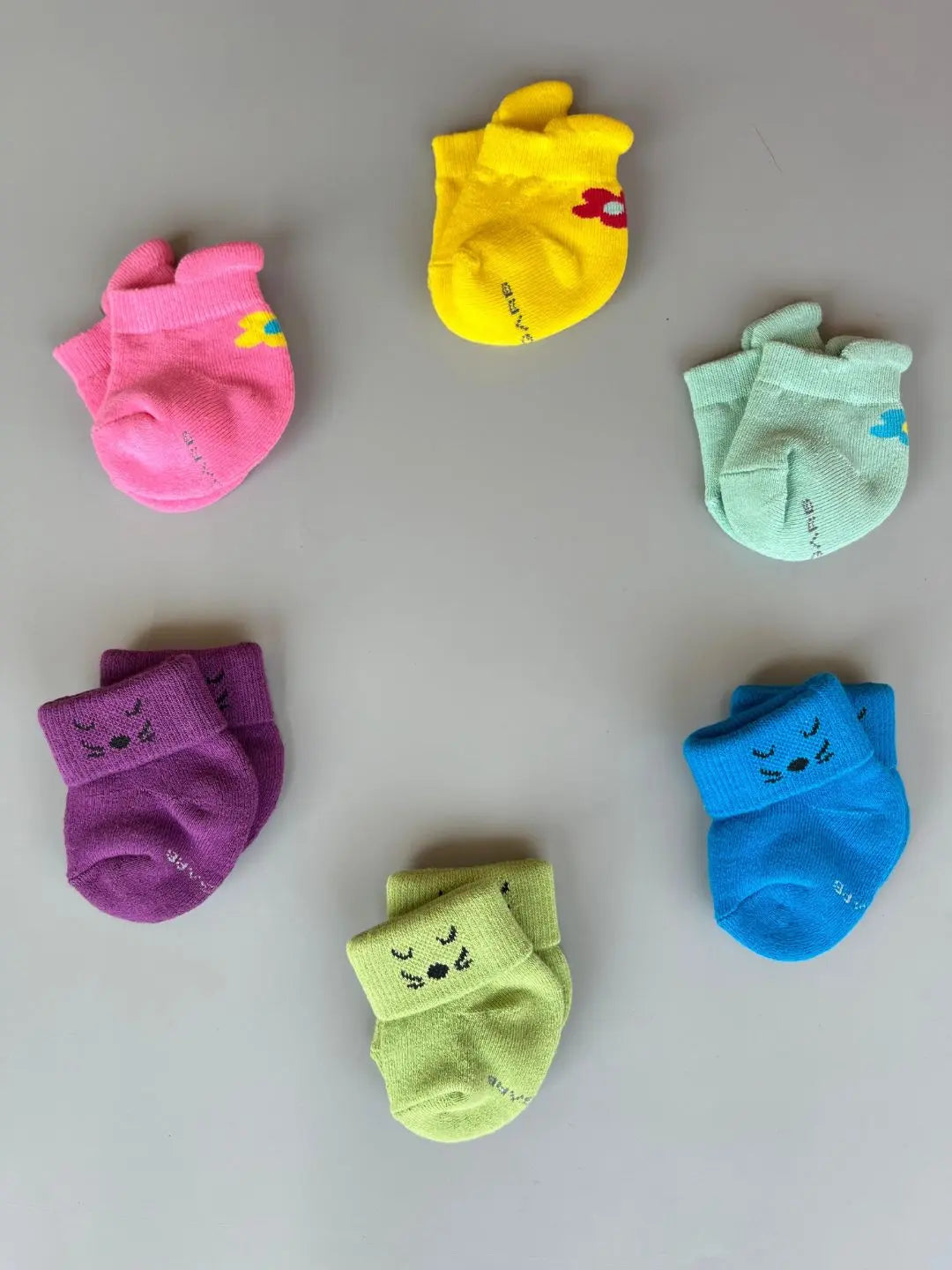 Air Garb brand baby combo socks for infants 0-1 years; a 6-pair collection arranged in a circle featuring three pairs with side flower designs (pink, yellow, mint) and three pairs with kitten face cuffs (purple, lime green, blue).